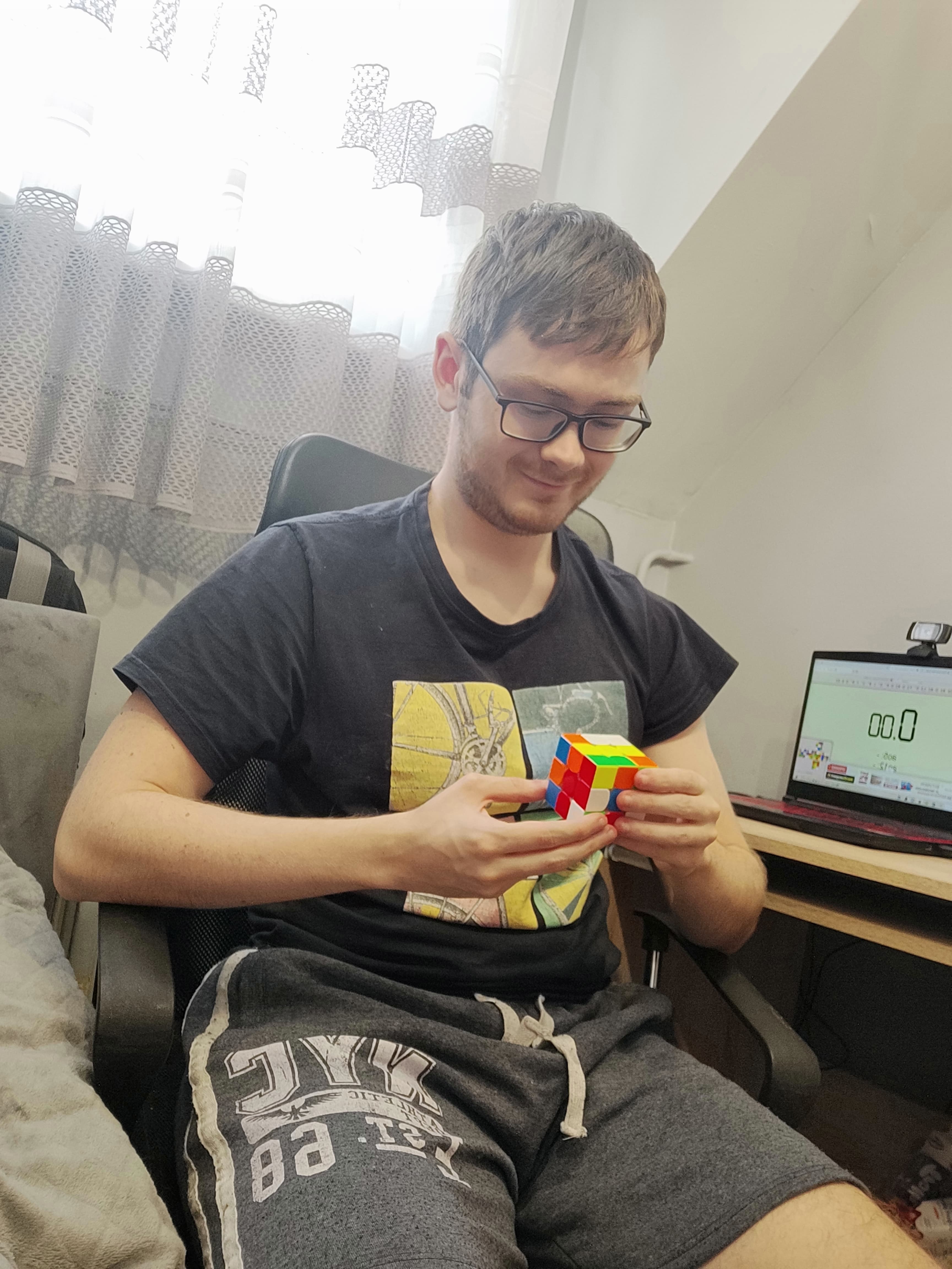 Mężczyzna układający kostkę Rubika, który siedzi na fotelu. Po prawej stoi biurko, a na nim laptop.